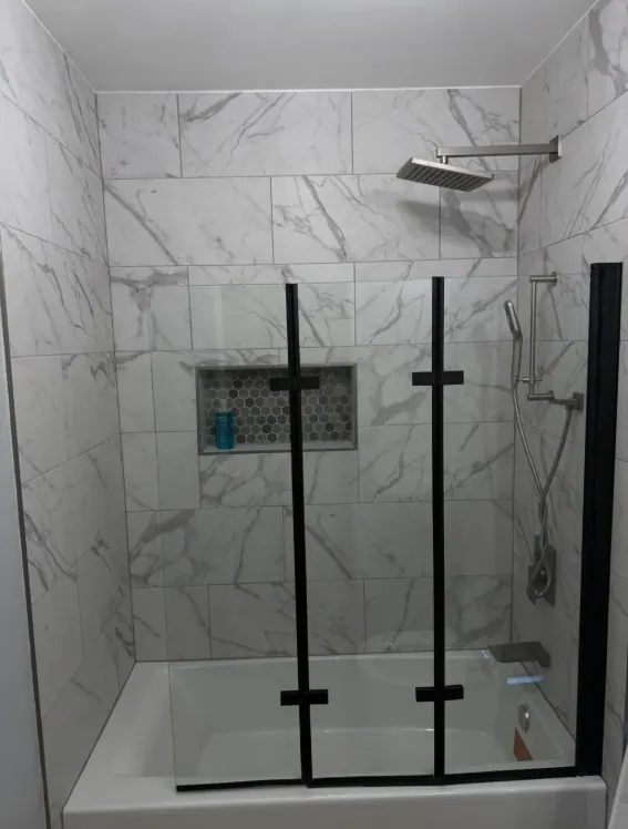 Tub surround in large marble-look tile with a hex mosaic niche and a black-framed folding glass screen
