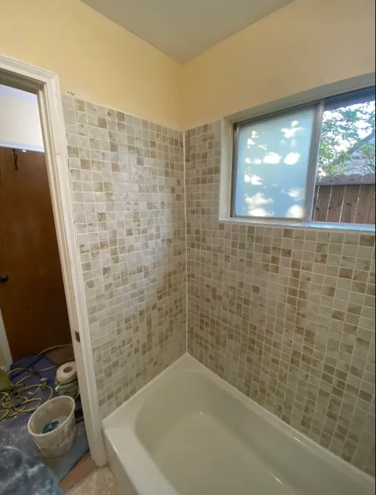 Beige square mosaic tub surround with the window wrapped in tile, work supplies still in the doorway