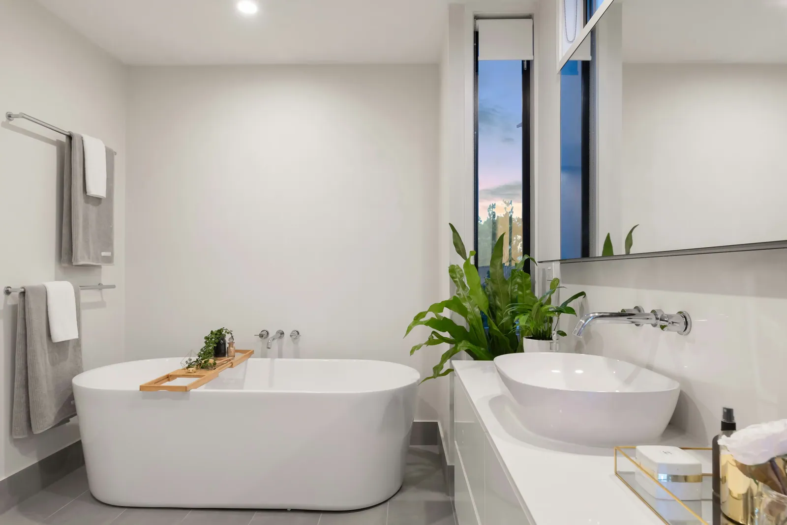 Well-lit bathroom with a freestanding white tub, gray tile floor, and long vanity