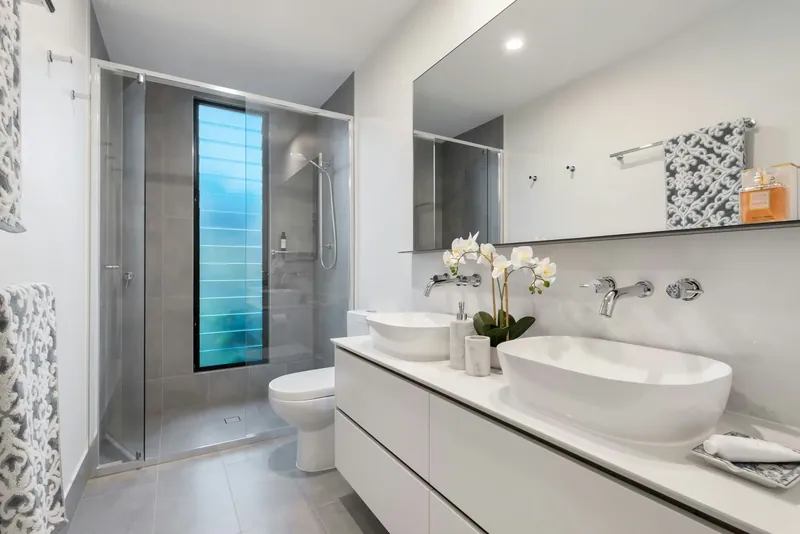 Double vessel-sink vanity facing a walk-in shower tiled in gray with a tall louvered window