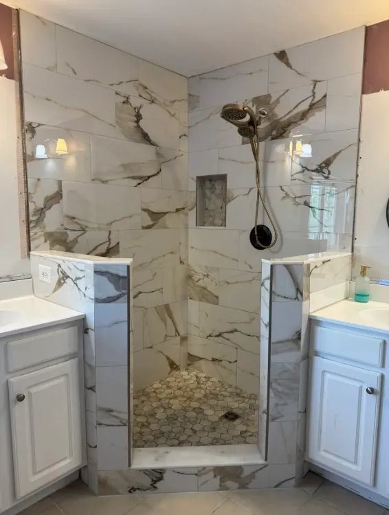 Open walk-in shower between two vanities, with warm-veined marble-look tile and a hex mosaic floor